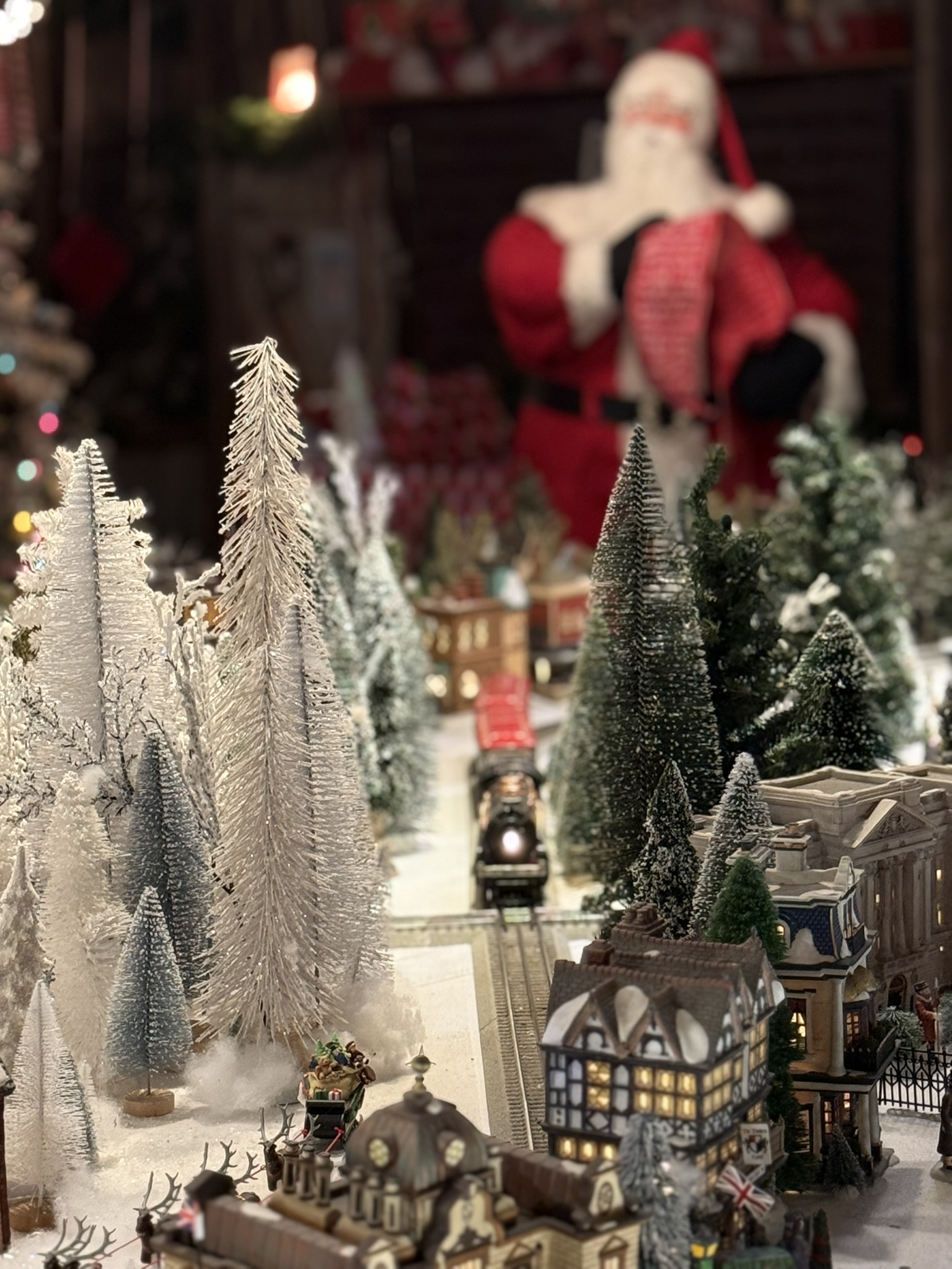 Christmas train village with a giant Santa figure looming warmly in the background, the locomotive's headlight visible between frosted bottle-brush trees